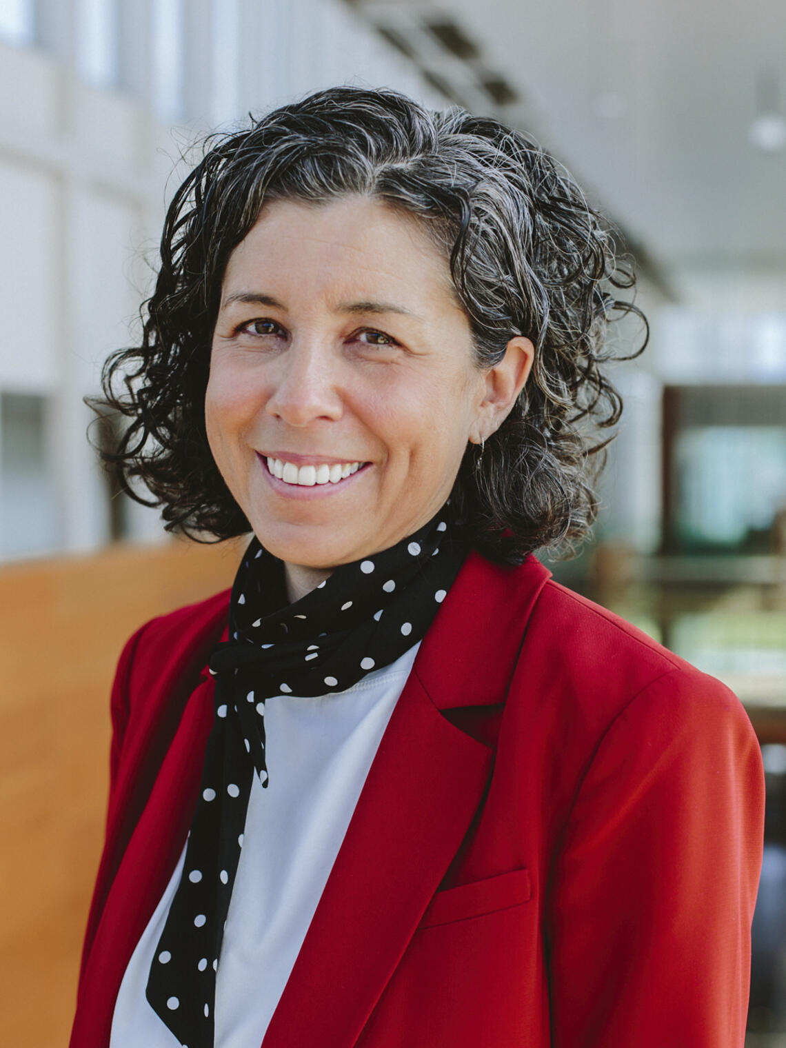 Leslie Reid, a white woman with dark curly hair, wearing a red blazer and a scarf, smiles inside the Taylor Institute for Teaching and Learning.