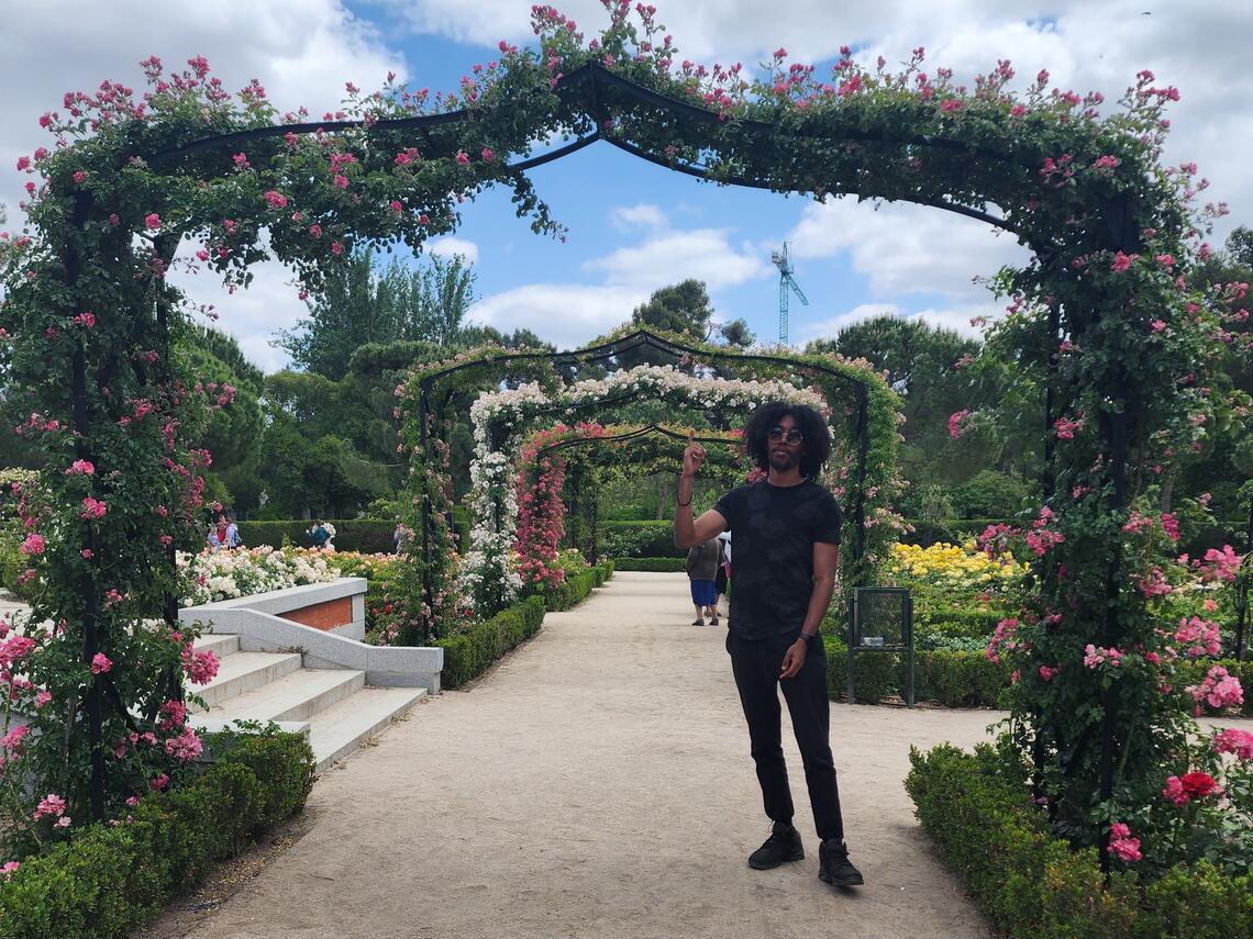 A young student with an afro stands on a garden pathway under an arch filled with plants.