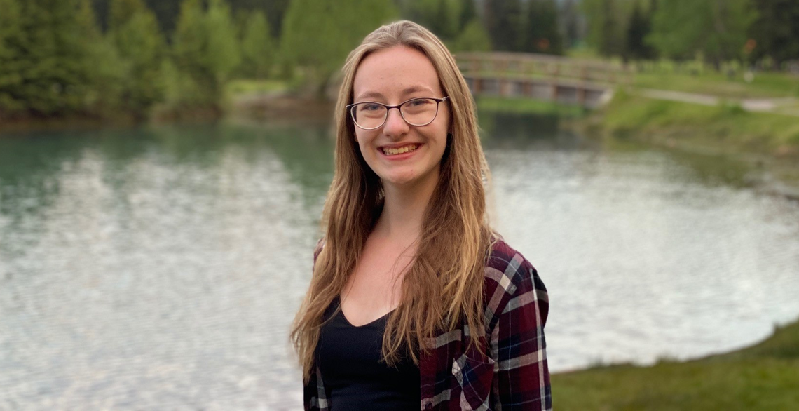 Young woman with shoulder length light brown hair wearing glasses with dark frames, a black top and a navy and burgundy plaid shirt, posing with a lake and mountains behind her