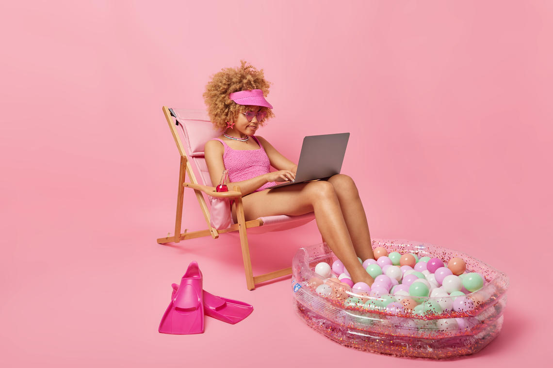 Female wearing all pink outfit works on a laptop while seated, surrounded by an assortment of summery items.