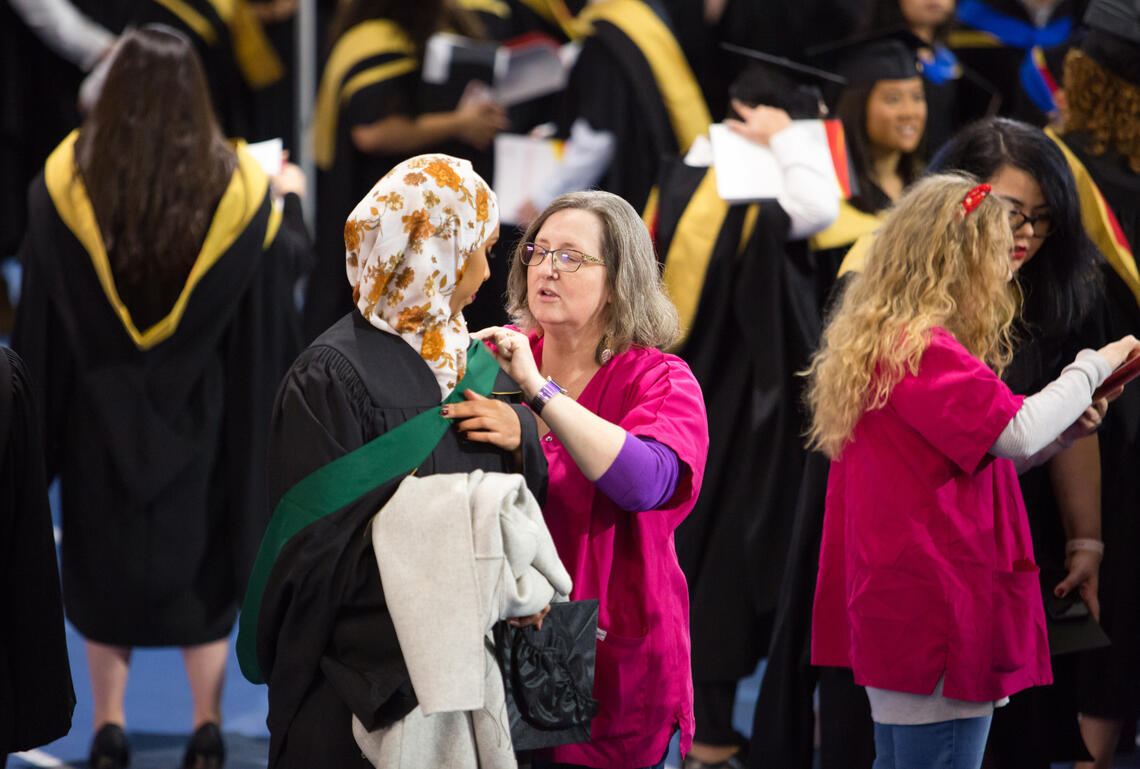 Two volunteers help graduates pin on gowns and provide directions as the grads get ready to walk the stage.