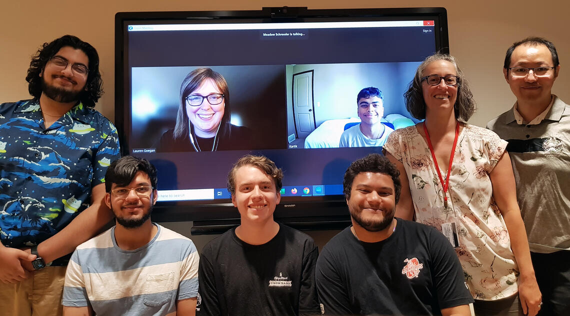 A group of people group around a zoom call screen while smiling at the camera
