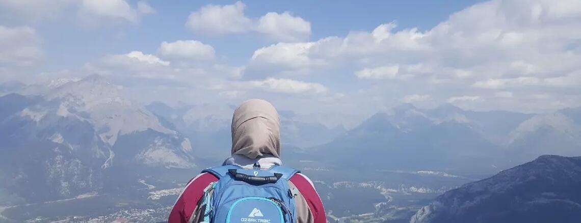 The back of a woman wearing a hijab and a backpack looking out over a mountain view.
