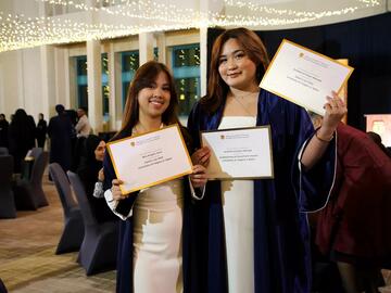 Two women smile at the camera holding their degrees