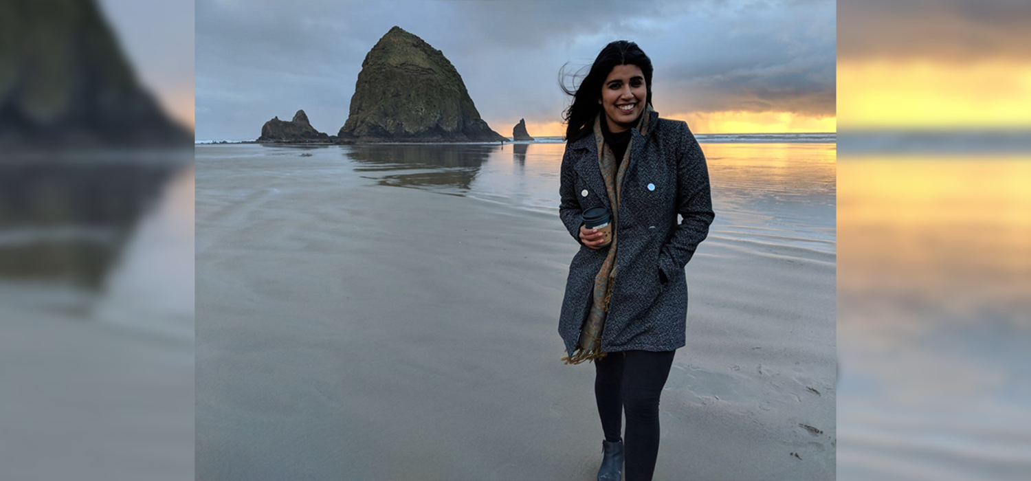 Alumna Tonie Minhas stands on a beach at sunset. She is wearing a jacket, holding a cup of coffee, and smiling at the camera. The sky is grey and gold behind her.