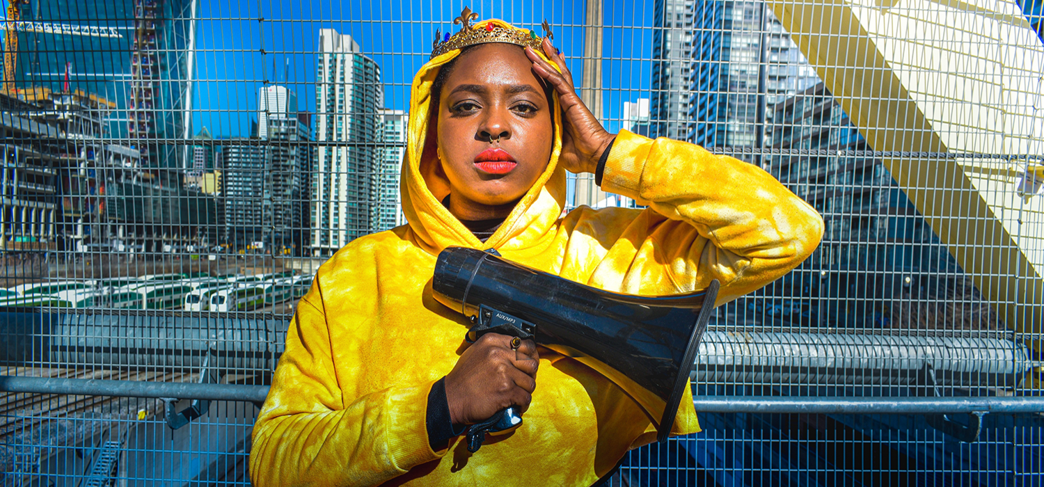 A Black woman wearing a crown and bright yellow hoodie stands against a wire fence. One hand is on her head, and the other holds a megaphone