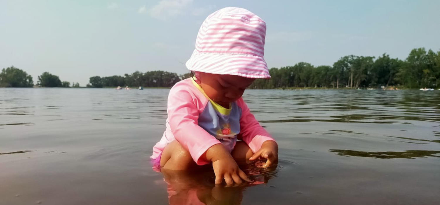A small child in a pink sun hat crouches in the shallow water of a lake.