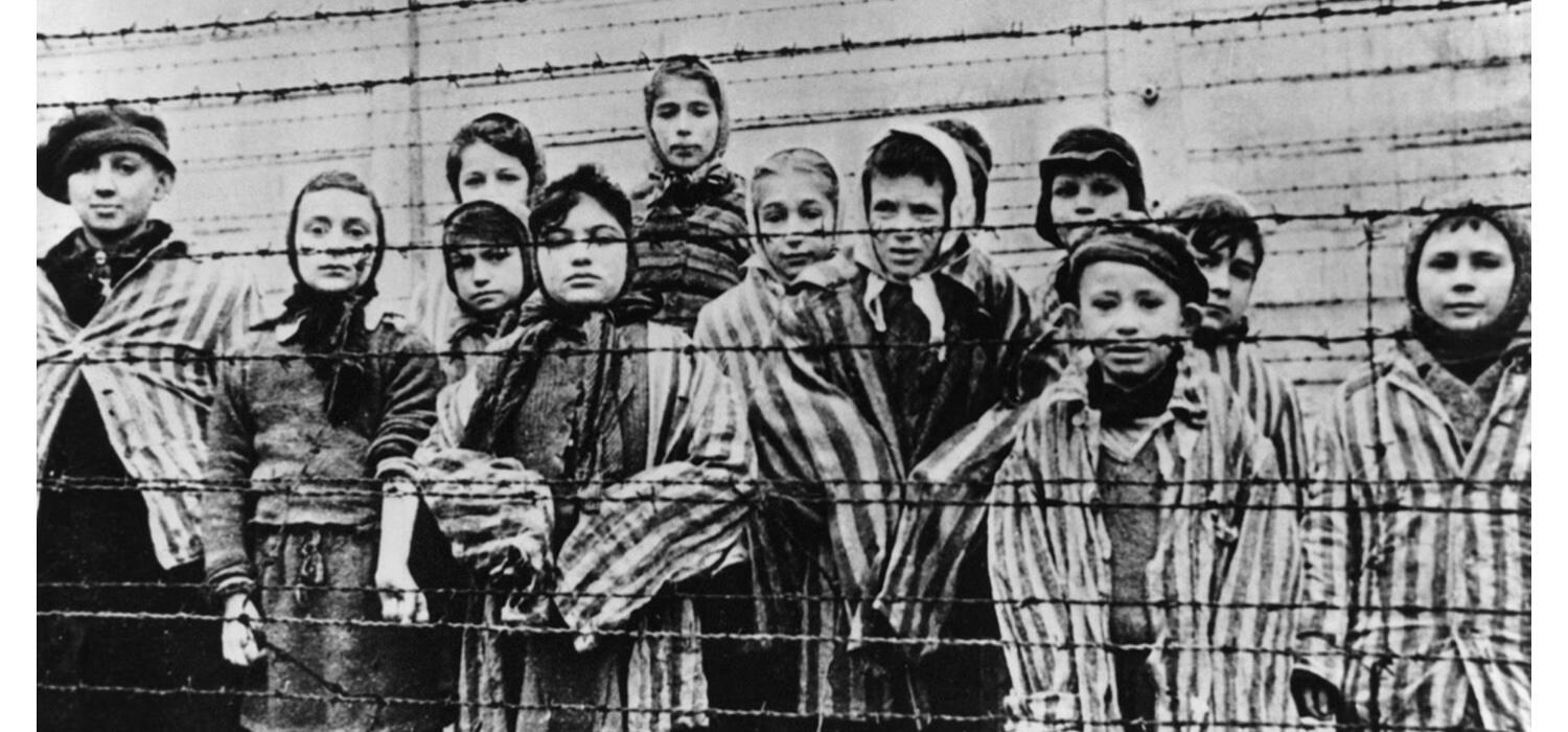 A black and white image depicting a group of children behind a barbed wire fence dressed in striped prison clothes at the Auschwitz concentration camp