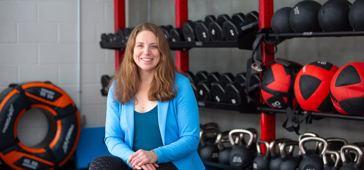 A woman is sitting in front of red and black gym equipment with a blue workout top and black pants on. She's smiling at the camera with her arms crossed over her knee and her long brown hair down.
