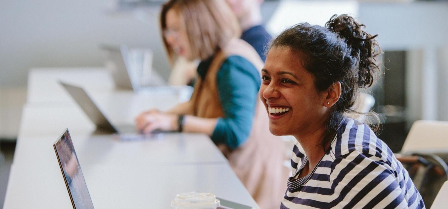 A woman with brown skin and brown hair in a ponytail, sits at a table with a laptop and is smiling. Behind her there are other people on laptops.