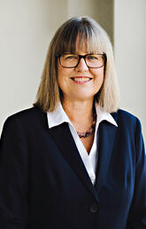 A woman wearing a button down and blazer and glasses smiles at the camera
