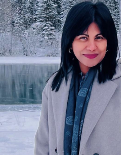 A woman stands smiling in front of wintery scene with snow covered fir trees and a frozen lake. She has dark hair, a blue scarf and a grey coat.