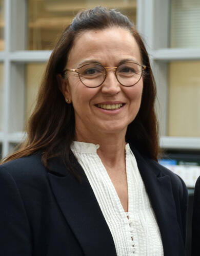 A woman with dark hair and glasses smiles at the camera in a dark suit jacket and whie blouse